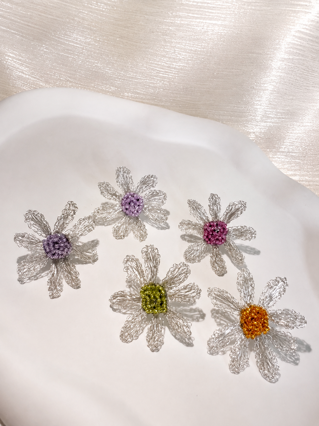 Five small daisy-shaped embellishments with textured clear petals and colorful centers in purple, pink, yellow, and orange, arranged on a white dish against a shimmering light background.