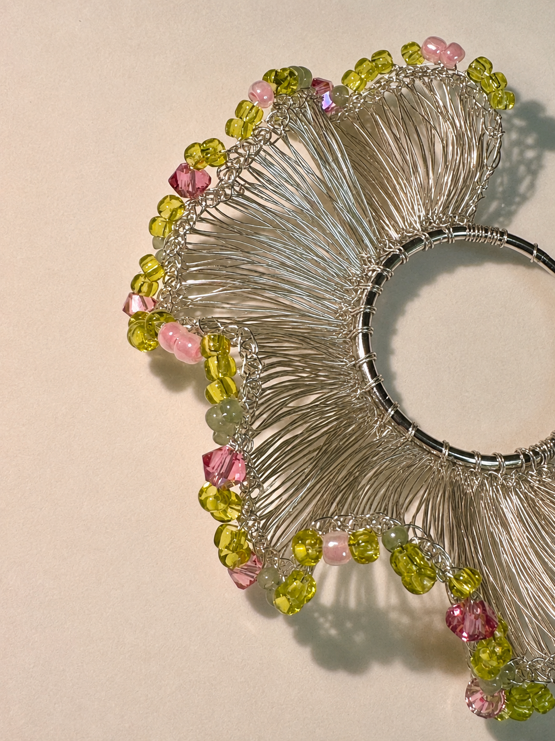 A close-up of a silver-toned wire collar necklace with faceted green and pink beads along the outer edge, photographed on a plain light beige background.
